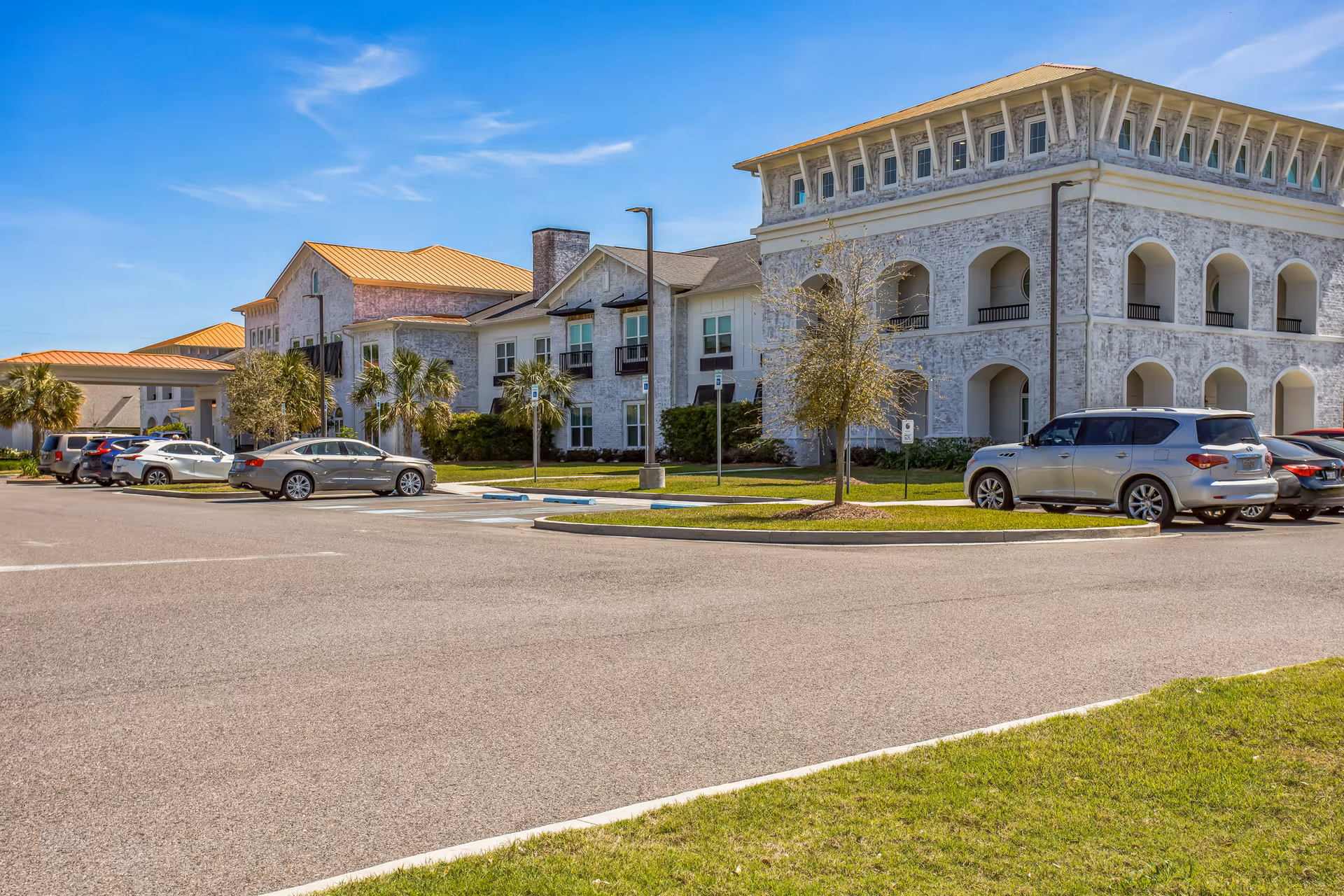 Exterior view of The Blake at Colonial Club senior living facility showing a large building with white textured walls, arched balconies, and a parking lot with several cars parked. The sky is clear and blue with some scattered clouds.