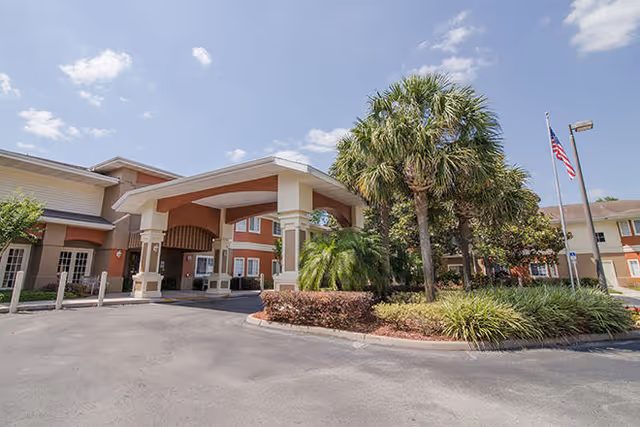 Exterior view of Brookdale Altamonte Springs senior living facility showing the main entrance with a covered drop-off area, palm trees, landscaping, and an American flag on a flagpole under a partly cloudy sky.