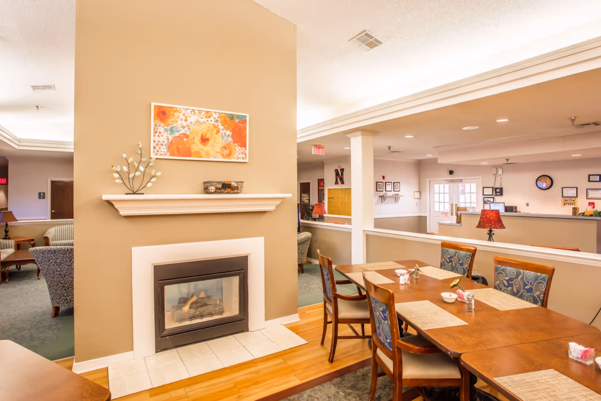 A cozy interior space featuring a dining table with six chairs, a beige wall with a modern fireplace, and a colorful floral painting above the mantel. The room has warm lighting, a mix of carpet and hardwood flooring, and a view into an adjacent area with more seating and a reception desk.