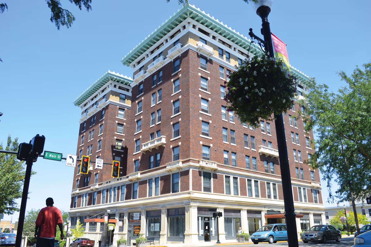 A multi-story brick building with decorative white and green trim on the roof, located at a street corner with traffic lights and street signs. There are cars parked along the street and a person walking on the sidewalk. Trees and a hanging flower basket on a street lamp are also visible.