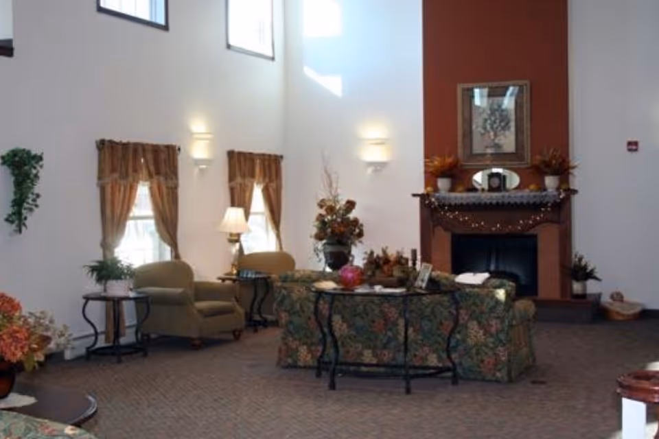 A cozy living room area with floral patterned sofas and armchairs, a fireplace with decorative plants and a framed picture above it, two windows with brown curtains, and several side tables with lamps and plants.