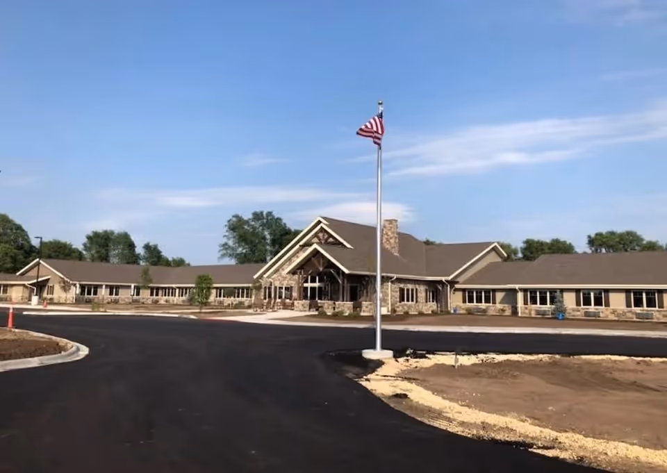 Exterior view of a single-story retirement living facility building with a stone facade and a pitched roof under a clear blue sky. An American flag is flying on a flagpole in front of the building, and there is a newly paved driveway and some landscaping in progress.