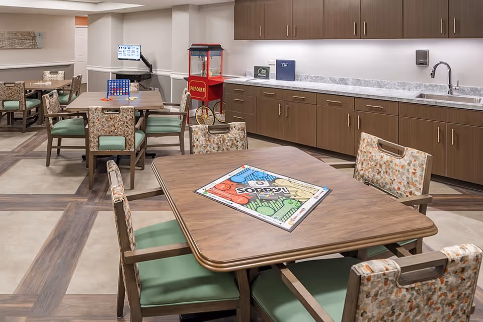 A well-lit common area with several tables and chairs, each table set up with board games like Sorry and Connect Four. The room features a long countertop with a sink and cabinets above and below. A red popcorn machine is visible in the background near a computer station.