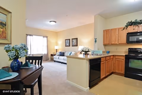 Interior view of a senior living facility showing a combined living room and kitchen area. The living room has a white sofa with blue and white cushions, two framed pictures on the wall, a side table with a lamp, and a window with curtains. The kitchen features wooden cabinets, a black dishwasher, a black stove, and a countertop with a flower arrangement. A dining table with chairs and a blue vase with flowers is visible in the foreground.