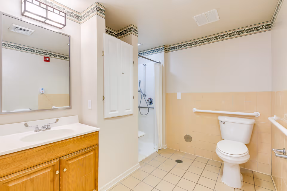A clean and accessible bathroom with beige tiled floor and walls, a wooden vanity with a white sink and faucet, a large mirror above the sink, a white toilet with grab bars on both sides, and a walk-in shower with a white curtain and a handheld showerhead.