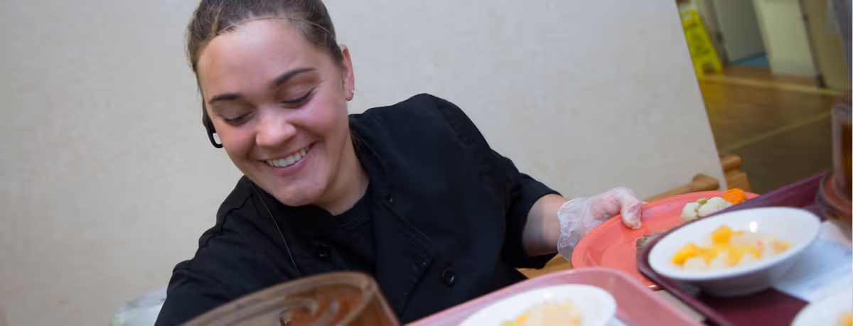 A smiling woman wearing a black uniform and a hairnet is serving food on trays with bowls of fruit salad in a dining or cafeteria setting.