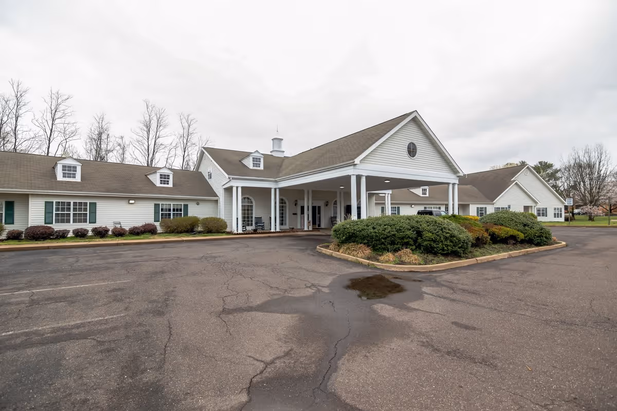 Exterior view of a single-story senior living facility building with white siding, a covered entrance supported by white columns, and a paved driveway with some bushes and trees around.