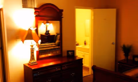 A warmly lit bedroom featuring a wooden dresser with a large mirror and a table lamp on top. The reflection in the mirror shows a cushioned chair. An open door reveals a bathroom with a sink and a fire extinguisher mounted on the wall. A small plant is visible on a piece of furniture to the right.