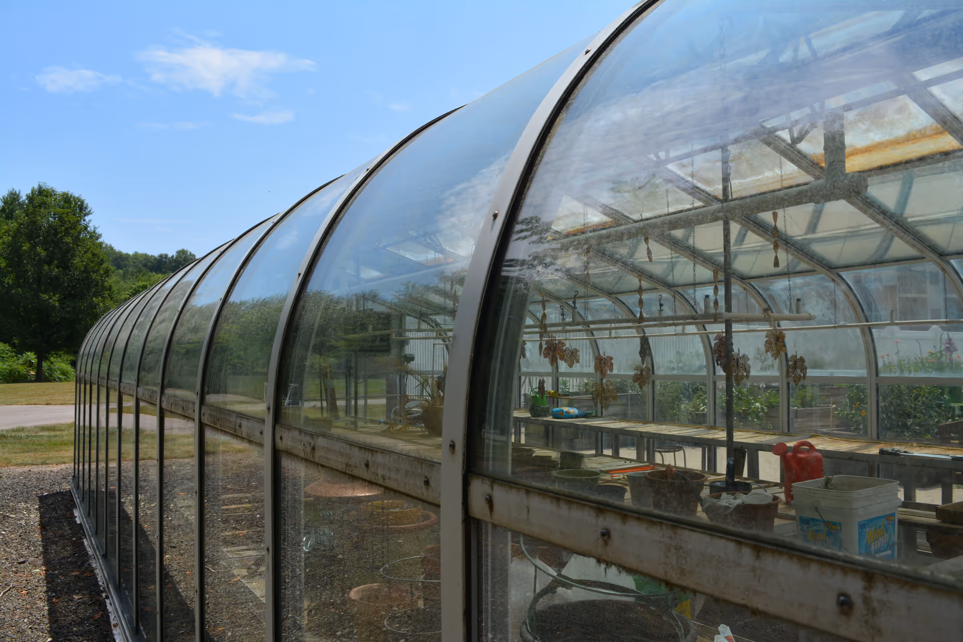 Exterior view of a long greenhouse structure with curved transparent panels. Inside, there are tables with gardening supplies, pots, and hanging dried plants. The greenhouse is situated outdoors with trees and grass visible in the background under a partly cloudy sky.