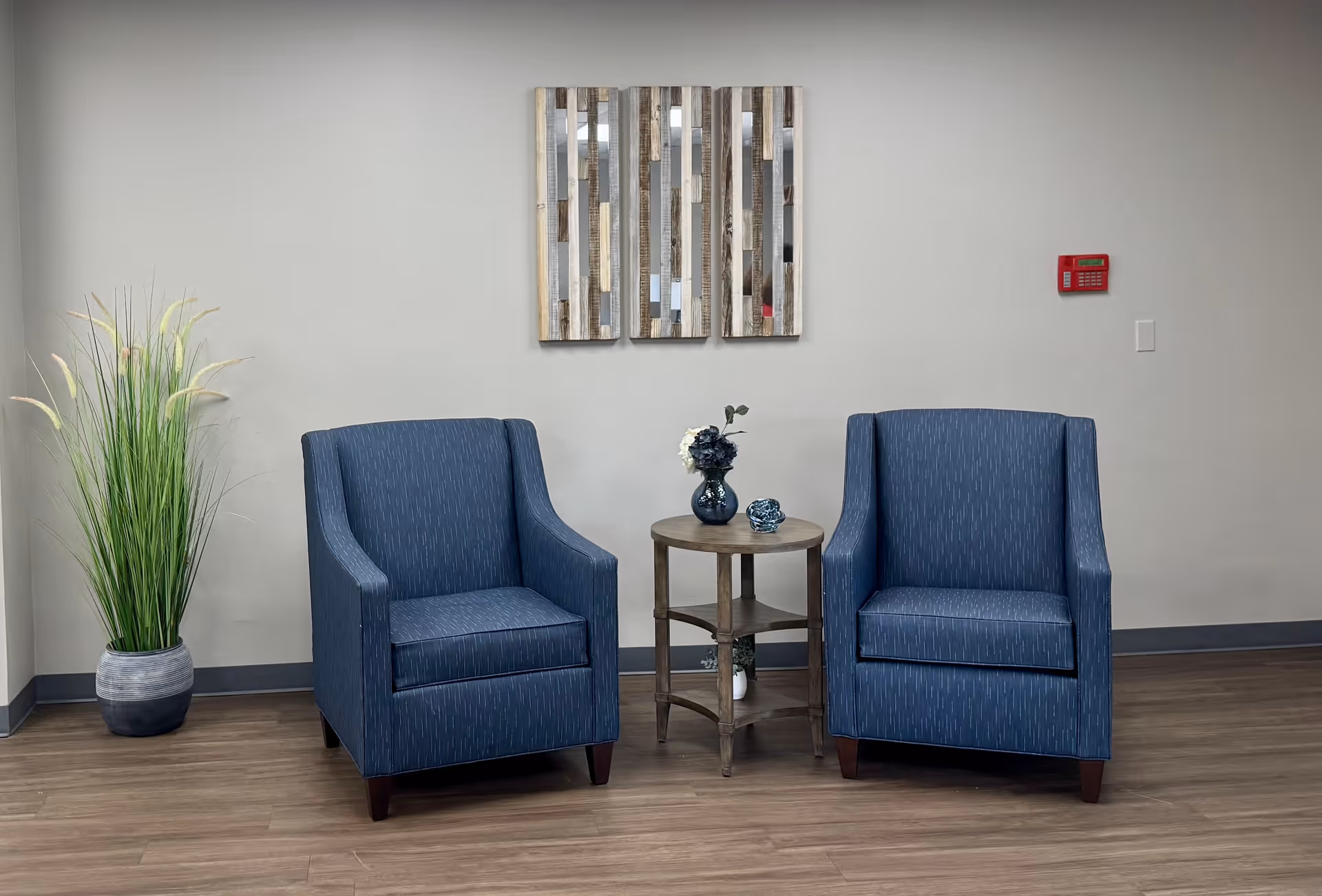 Two blue upholstered armchairs with a small round wooden table between them, decorated with a vase of flowers and a small decorative item. A tall potted plant is placed to the left, and a three-panel wooden wall art hangs on the light gray wall behind the chairs. A red fire alarm is mounted on the wall to the right.