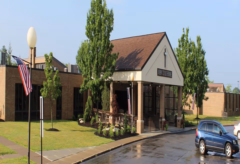 Exterior view of Saint John XXIII Home, a brick building with a peaked roof entrance featuring a cross and a sign. There are trees and American flags around the building, with a wet driveway and parked cars in front.