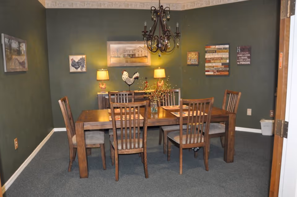 A dining room with a wooden table and six chairs on a carpeted floor. The walls are painted green and decorated with framed pictures and wall art. Two table lamps with rooster-shaped bases are on a sideboard against the back wall, and a chandelier hangs above the table. A vase with flowers is placed in the center of the table.