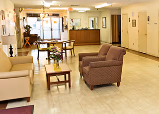 A spacious healthcare facility lobby with beige and brown armchairs, a wooden coffee table with a flower arrangement, and a reception desk in the background. The floor is tiled, and the walls are decorated with framed pictures. There is a glass door entrance at the far end.