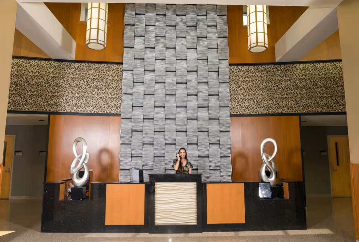 Reception area of Elmwood Hills Healthcare Center with a woman sitting behind a modern desk. The background features a tall textured wall panel and two abstract silver sculptures on either side of the desk. Warm lighting fixtures hang from the ceiling.