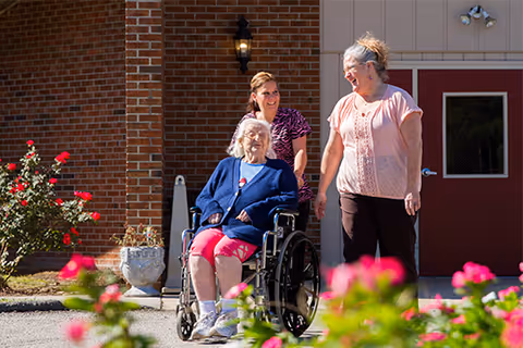 An elderly woman in a wheelchair is being pushed by a caregiver outside a brick building. Another woman walks alongside them, smiling. There are pink flowers in the foreground and a lantern mounted on the wall near the entrance door behind them.
