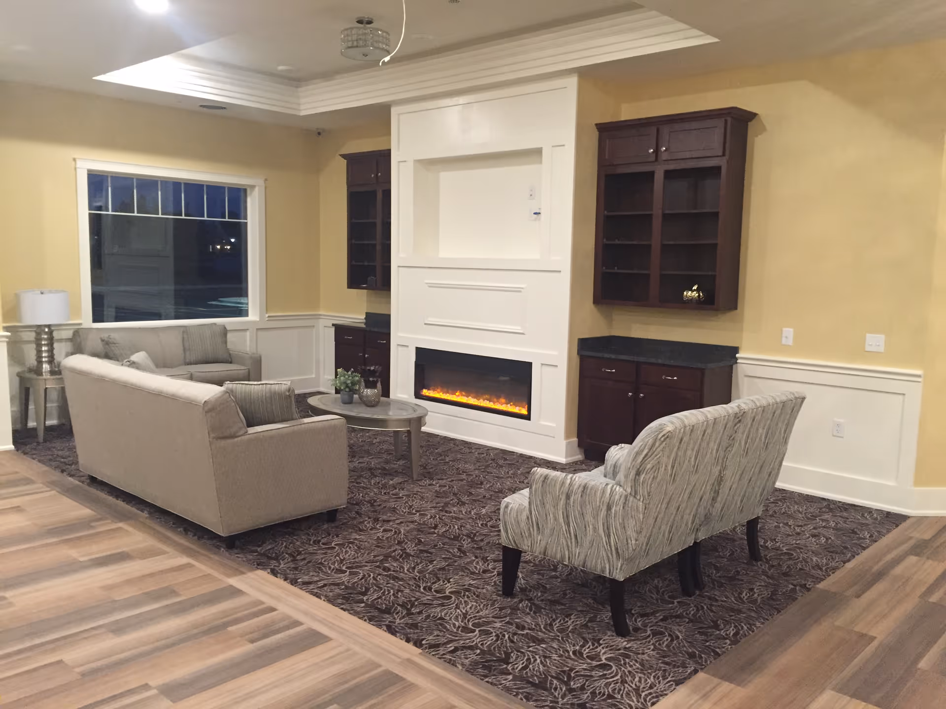 A cozy living room area with beige walls and a patterned carpet. The room features a beige sofa, a matching loveseat, and a patterned armchair arranged around a small oval coffee table. There is a modern electric fireplace built into a white wall unit with dark wood cabinets on either side. A large window is visible on the left side, and a floor lamp stands next to the sofa.