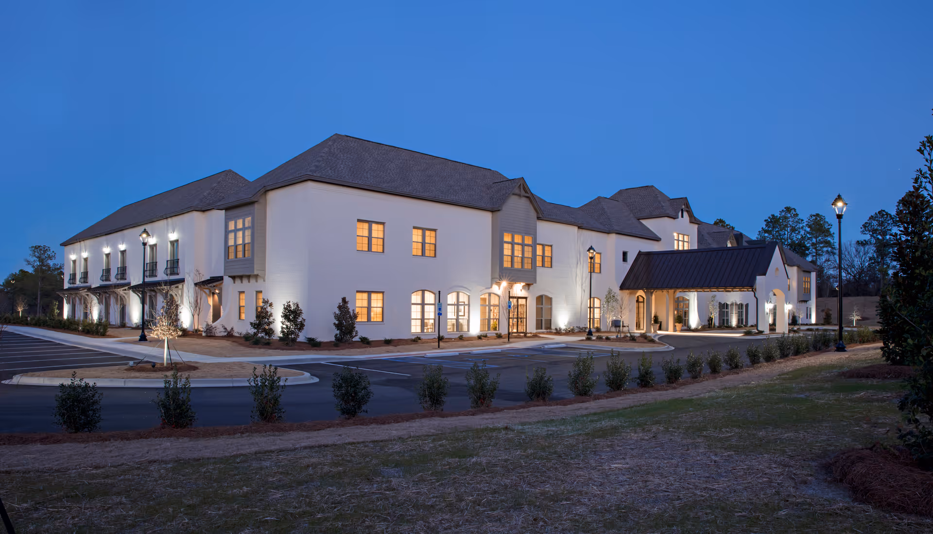 Illuminated two-story senior living building at dusk with a covered entrance and adjacent parking area.