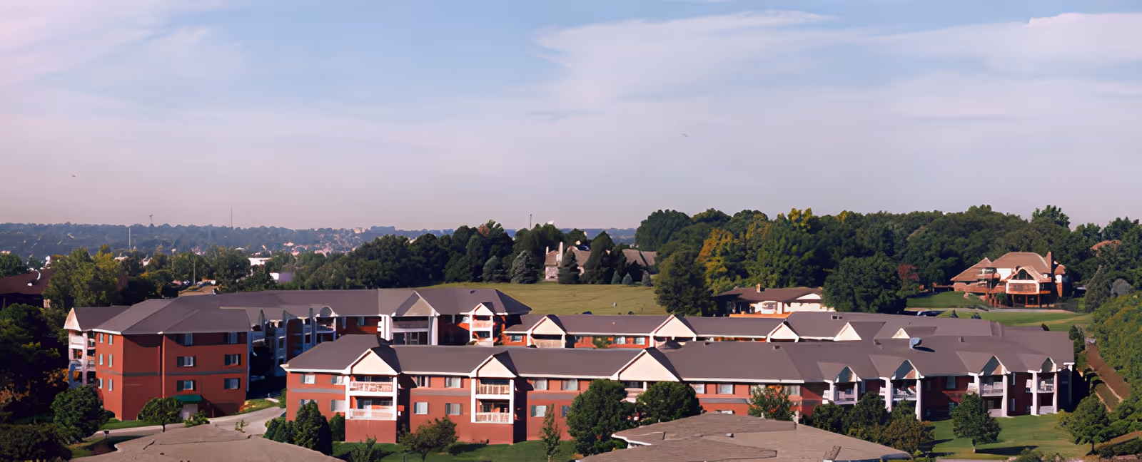 A large senior living facility named Bloomfield with multiple connected buildings featuring red brick walls and gray roofs, surrounded by green lawns and trees under a partly cloudy sky.