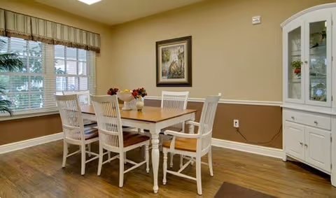 Dining room featuring a wooden table with six white chairs, a corner china cabinet, framed art, and a window with blinds.