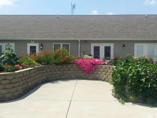 Outdoor courtyard area with a curved stone retaining wall filled with colorful flowers and plants, in front of a single-story building with multiple doors and windows under a clear blue sky.