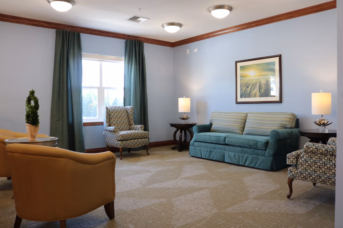 A cozy living room with light blue walls, a teal sofa with striped cushions, two patterned armchairs, two beige armchairs, a window with green curtains, two side tables with lamps, and a framed picture of a sunset over a field on the wall.