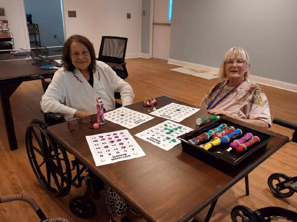 Two elderly women sitting at a wooden table in a room with light-colored walls and wooden flooring. Both women are in wheelchairs and appear to be playing bingo, with bingo cards and colorful bingo daubers on the table in front of them. One woman is wearing a white cardigan and the other a light pink floral top.
