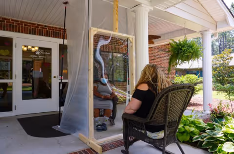Two women sitting outside the entrance of a brick building, separated by a clear plastic barrier mounted on a wooden frame. One woman is wearing a face mask and sitting on a chair inside the barrier, while the other woman sits on a wicker chair outside. The entrance has white columns and a hanging fern plant nearby.