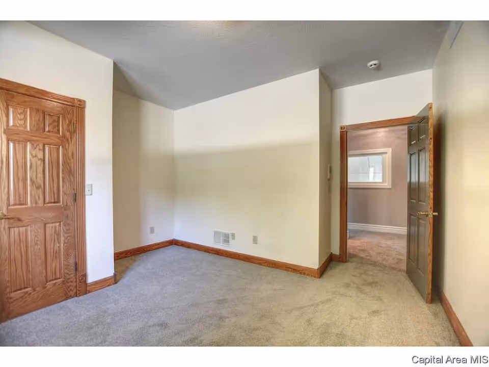 Empty room with beige walls, carpeted floor, wooden baseboards, and two wooden doors, one closed and one open leading to another room with a window.