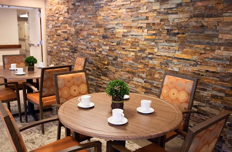 A cozy dining area with round wooden tables set with white cups and saucers. Each table has a small green plant centerpiece. The chairs have orange patterned cushions, and the background features a textured stone wall.