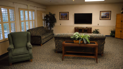 A cozy common living room with sofas, an armchair, a coffee table with plants, a wall-mounted TV, framed pictures, and large shuttered windows.