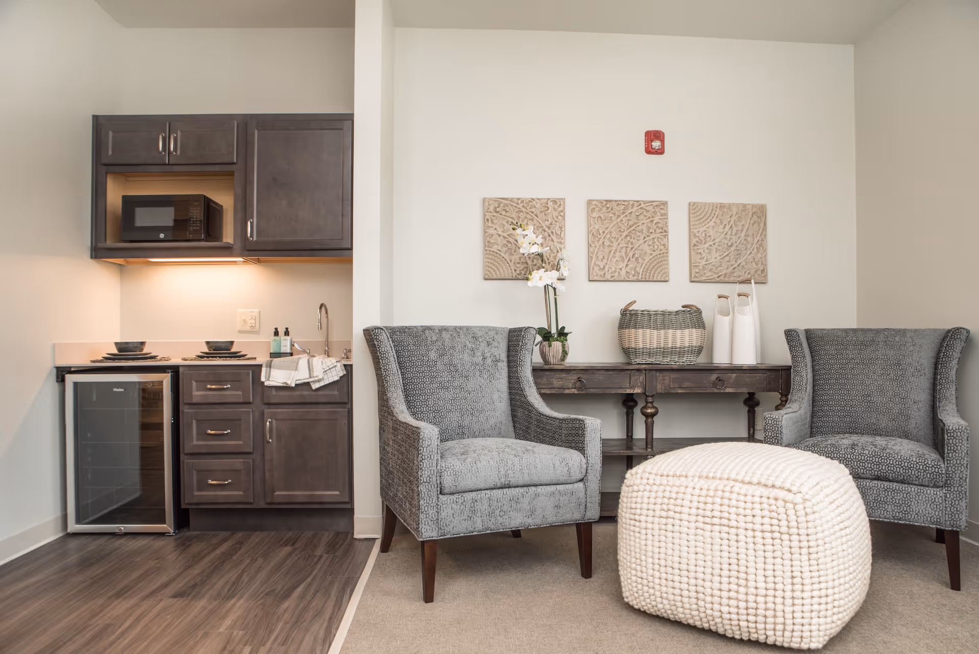 A cozy senior living area featuring two gray upholstered armchairs, a textured white ottoman, and a dark wooden console table with decorative items including a basket, two white vases, and a potted orchid. To the left, there is a small kitchenette with dark cabinets, a microwave, a mini fridge, a sink, and two place settings on the counter.