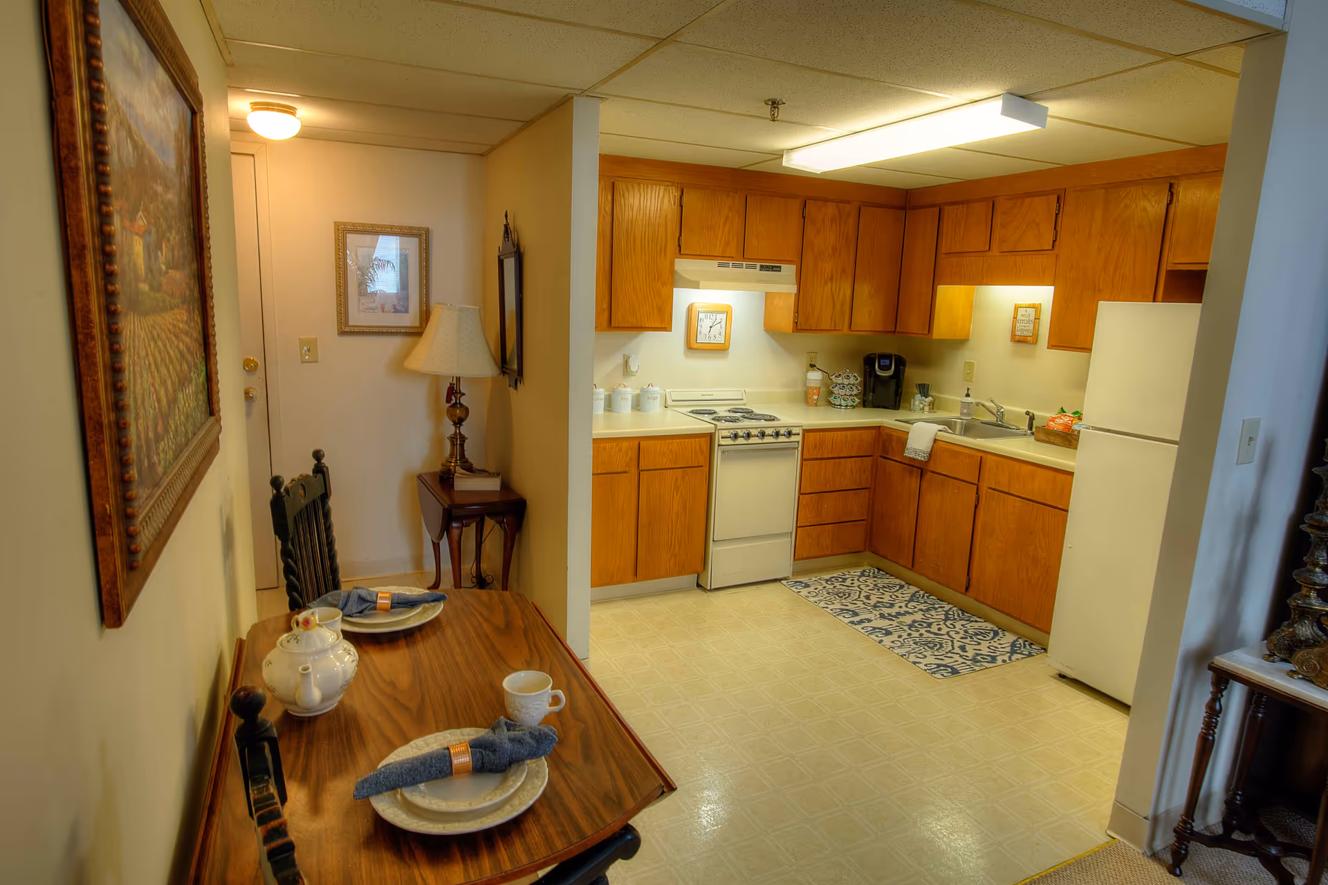 Small kitchenette with wooden cabinets, white appliances and a small dining table set for two.