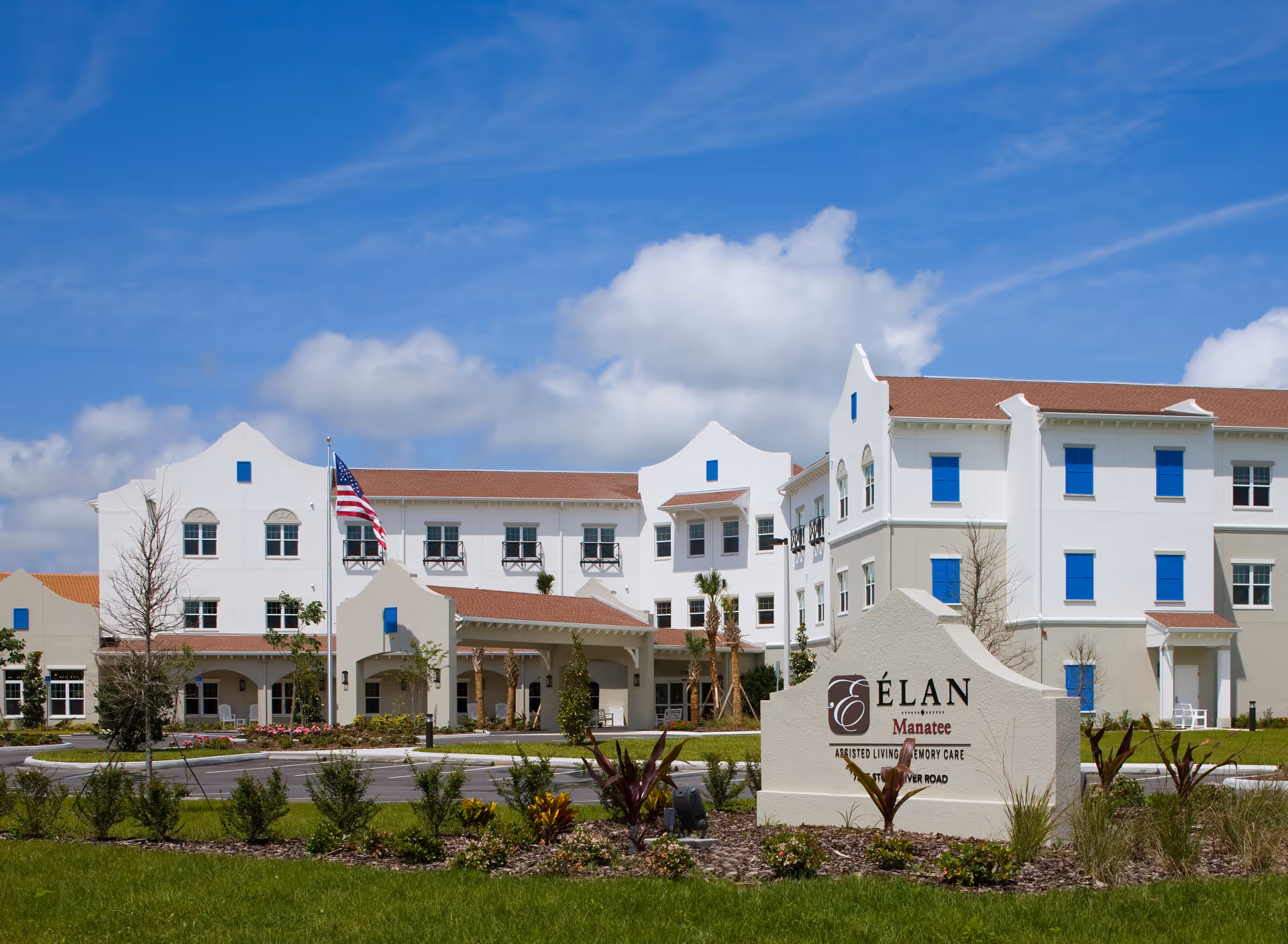 Exterior view of Élan Manatee, a multi-story assisted living and memory care facility with white and beige walls, blue window shutters, a red-tiled roof, and an American flag in front. The building is surrounded by landscaped greenery and a clear blue sky with some clouds.