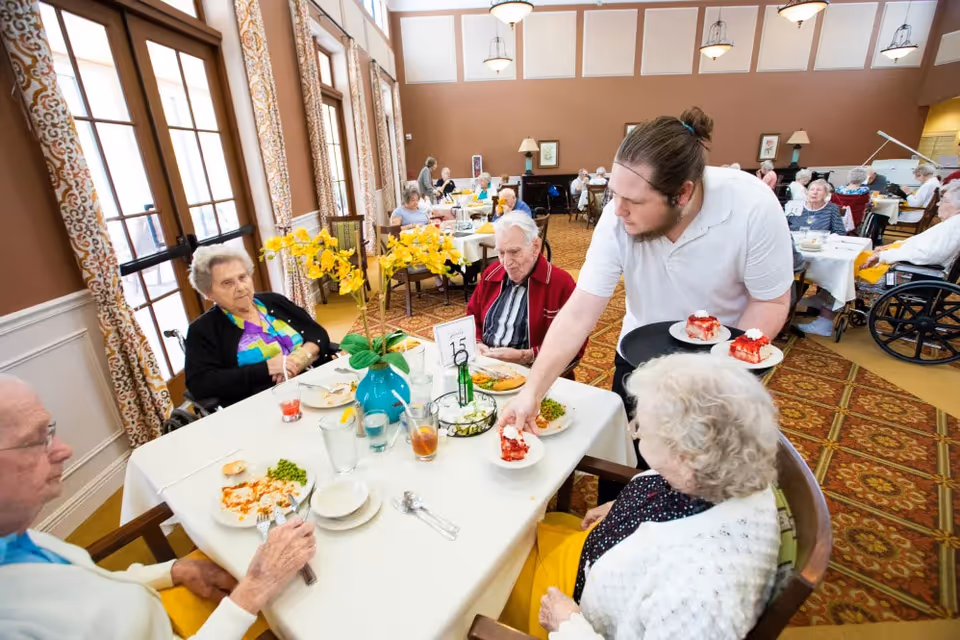 A staff member serves dessert to elderly residents seated around a table in a bright communal dining room.