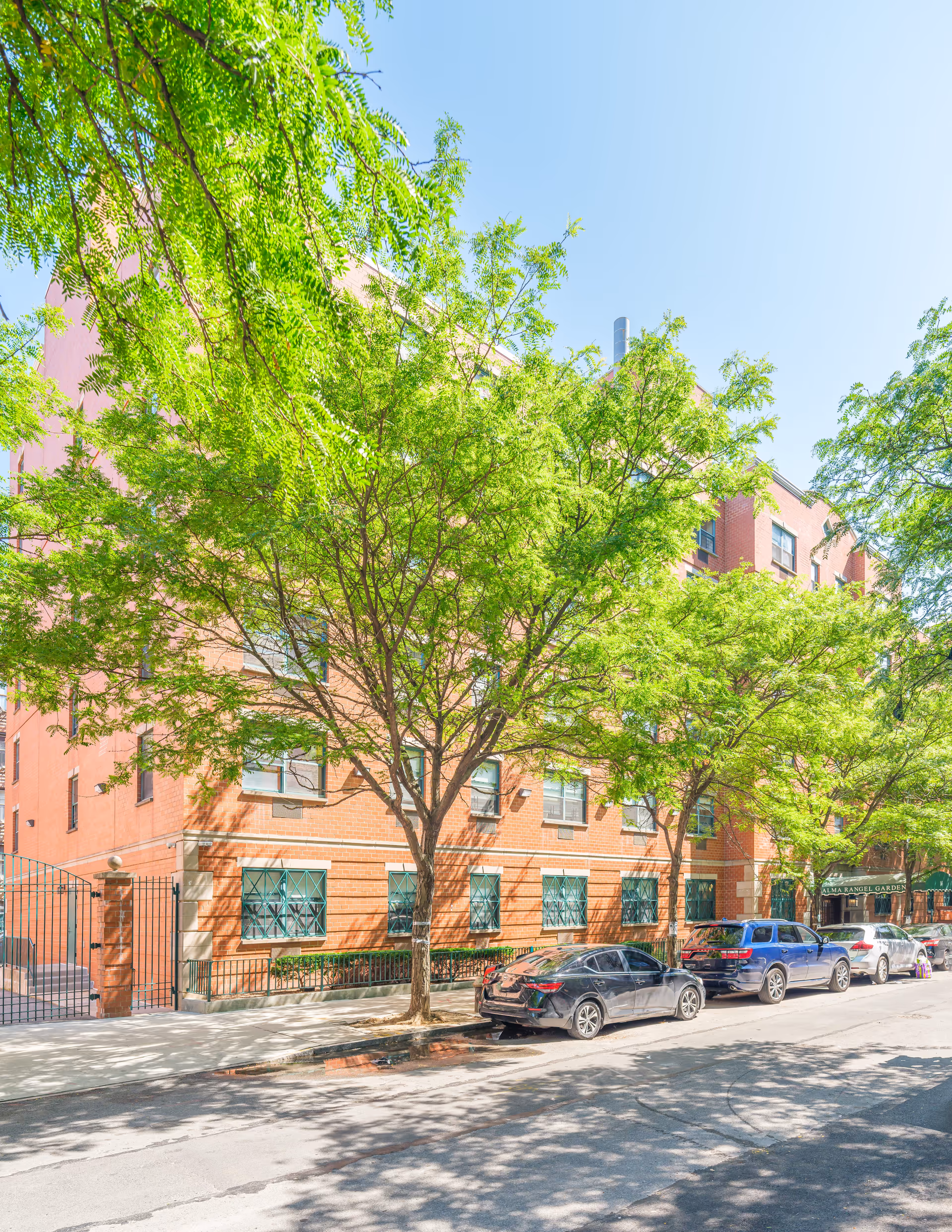 Street view of a red brick building partially obscured by green leafy trees. Several cars are parked along the curb in front of the building. A green awning with the text 'Alma Rangel Garden' is visible near the entrance.