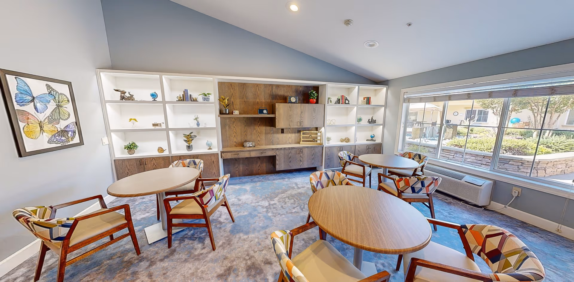 A bright and spacious common area with round wooden tables and colorful cushioned chairs. The room features a large window overlooking a garden area outside, built-in white shelves with decorative items, and a framed butterfly artwork on the wall.