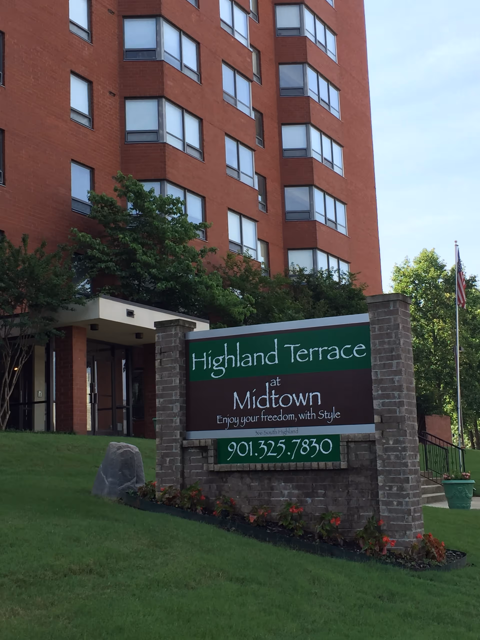 Exterior view of a multi-story red brick building with many windows, surrounded by greenery. In the foreground, there is a brick sign with green and brown panels that reads 'Highland Terrace at Midtown Enjoy your freedom, with Style' along with a phone number 901.325.7830. There is an American flag on a flagpole to the right of the sign.