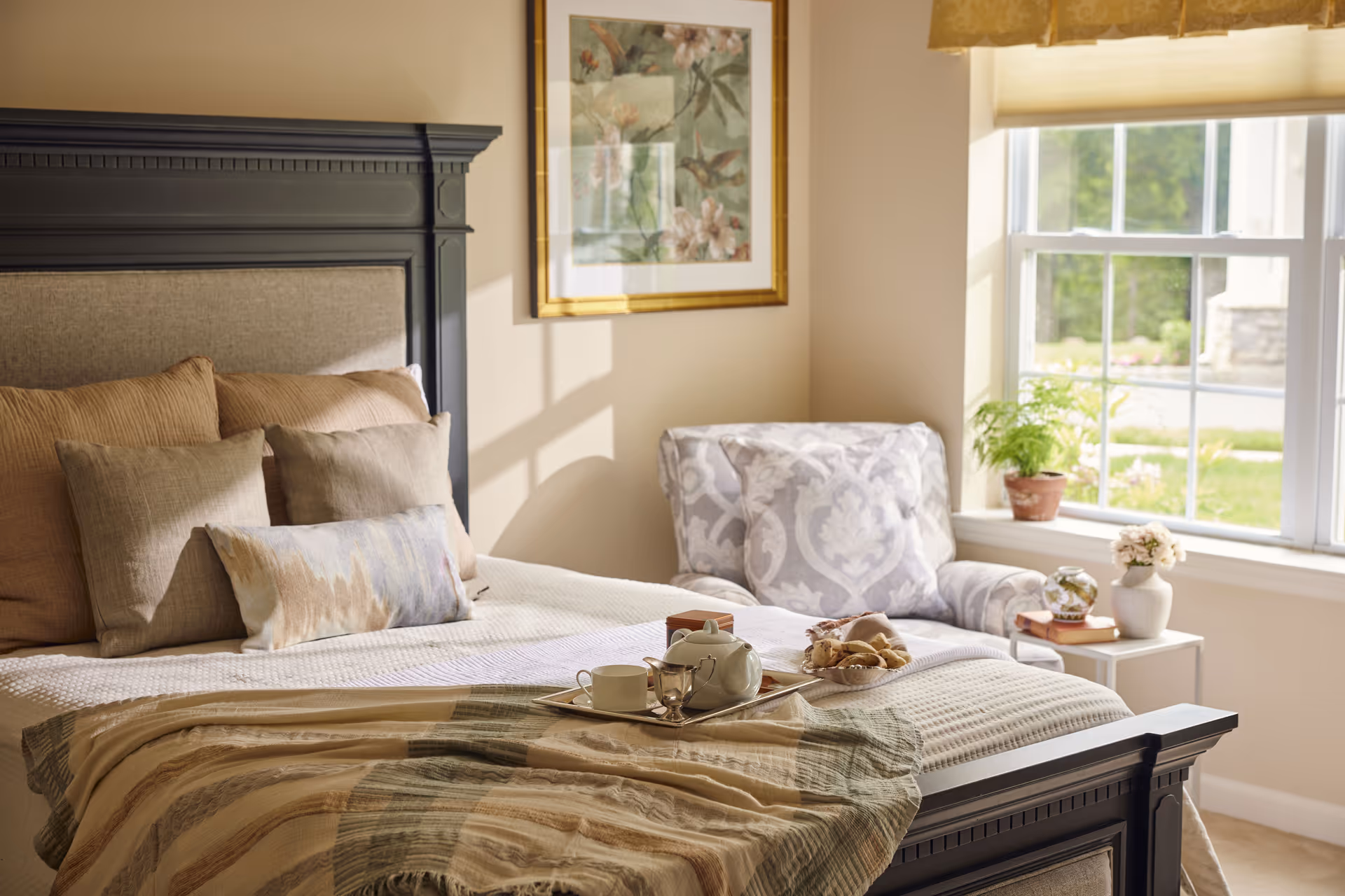 A cozy bedroom with a large bed featuring multiple pillows and a beige and green striped blanket. A tray with a teapot, cup, and snacks is placed on the bed. Next to the bed is a patterned armchair near a window with a view of greenery outside. A small side table holds a potted plant, a vase with flowers, and some books. A framed floral artwork hangs on the wall above the armchair.