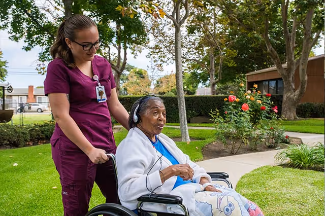 A caregiver in a purple uniform is pushing an elderly woman in a wheelchair along a paved path in a garden area. The elderly woman is wearing headphones and a white jacket, and there are trees, bushes, and flowers around them with a building in the background.