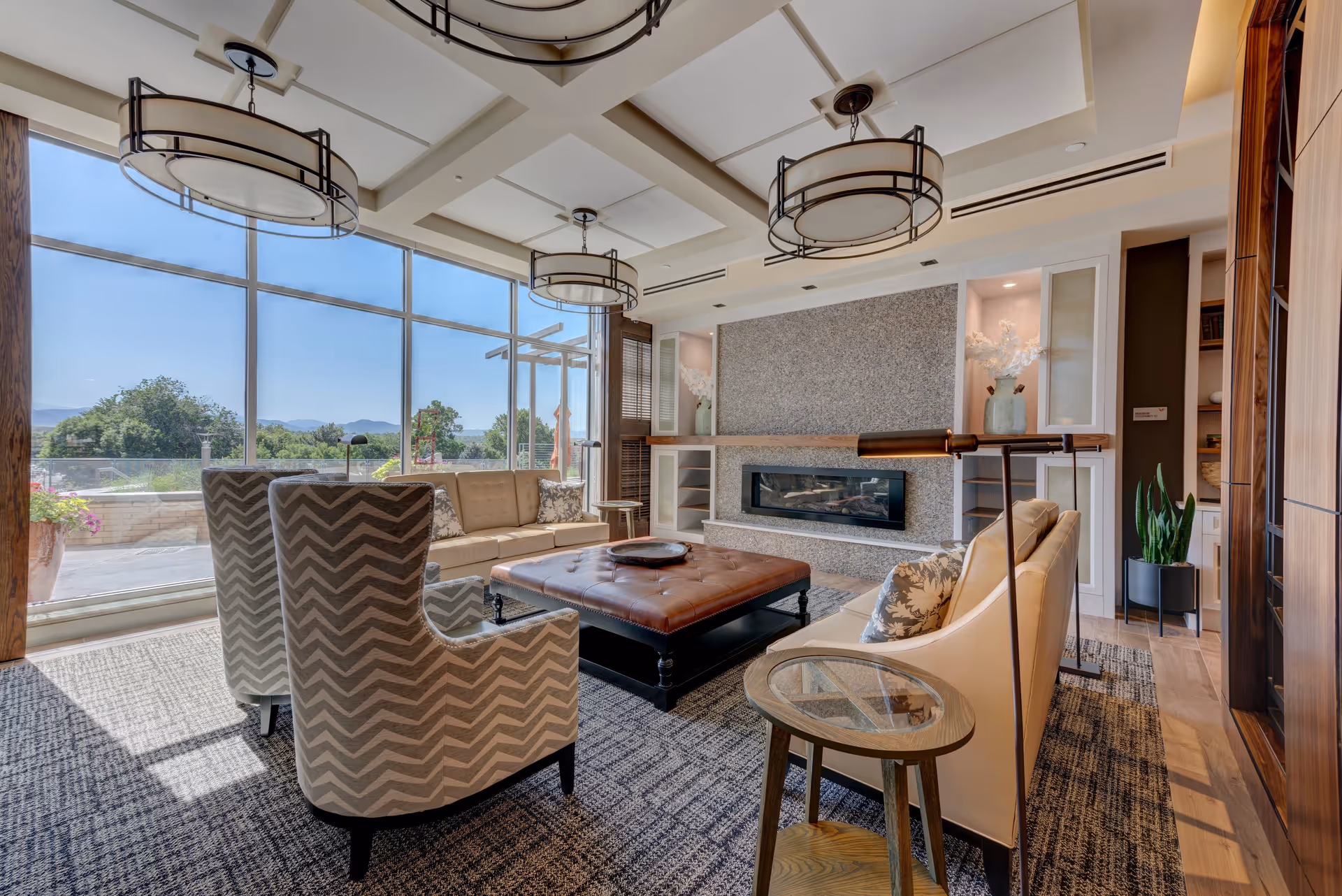 A bright and modern living room with large floor-to-ceiling windows showing an outdoor view. The room features a beige sofa, two patterned armchairs, a large square leather ottoman coffee table, a fireplace set in a stone wall, and contemporary ceiling light fixtures. There are decorative plants and shelves on either side of the fireplace.