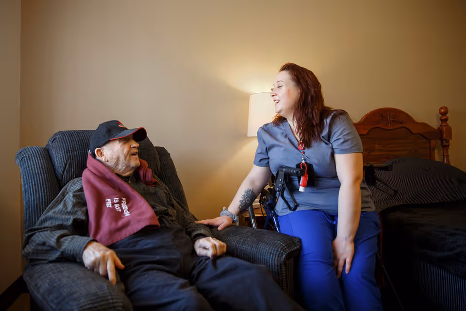A caregiver sits beside an elderly man in a recliner in a bedroom, smiling as they hold hands.