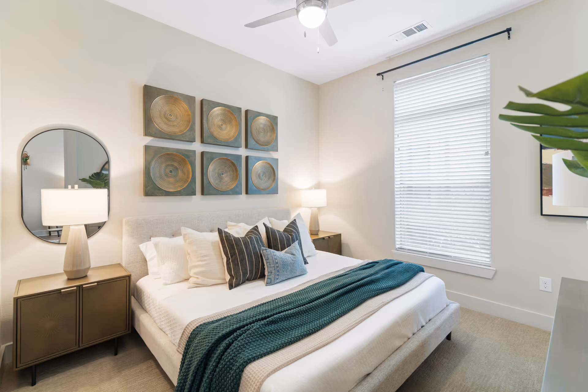 A bright and cozy bedroom with a large bed featuring white and blue pillows and a teal knitted throw blanket. There are two bedside tables with lamps, a round mirror on the left wall, six decorative circular art pieces above the bed, a window with white blinds, and a ceiling fan with a light.