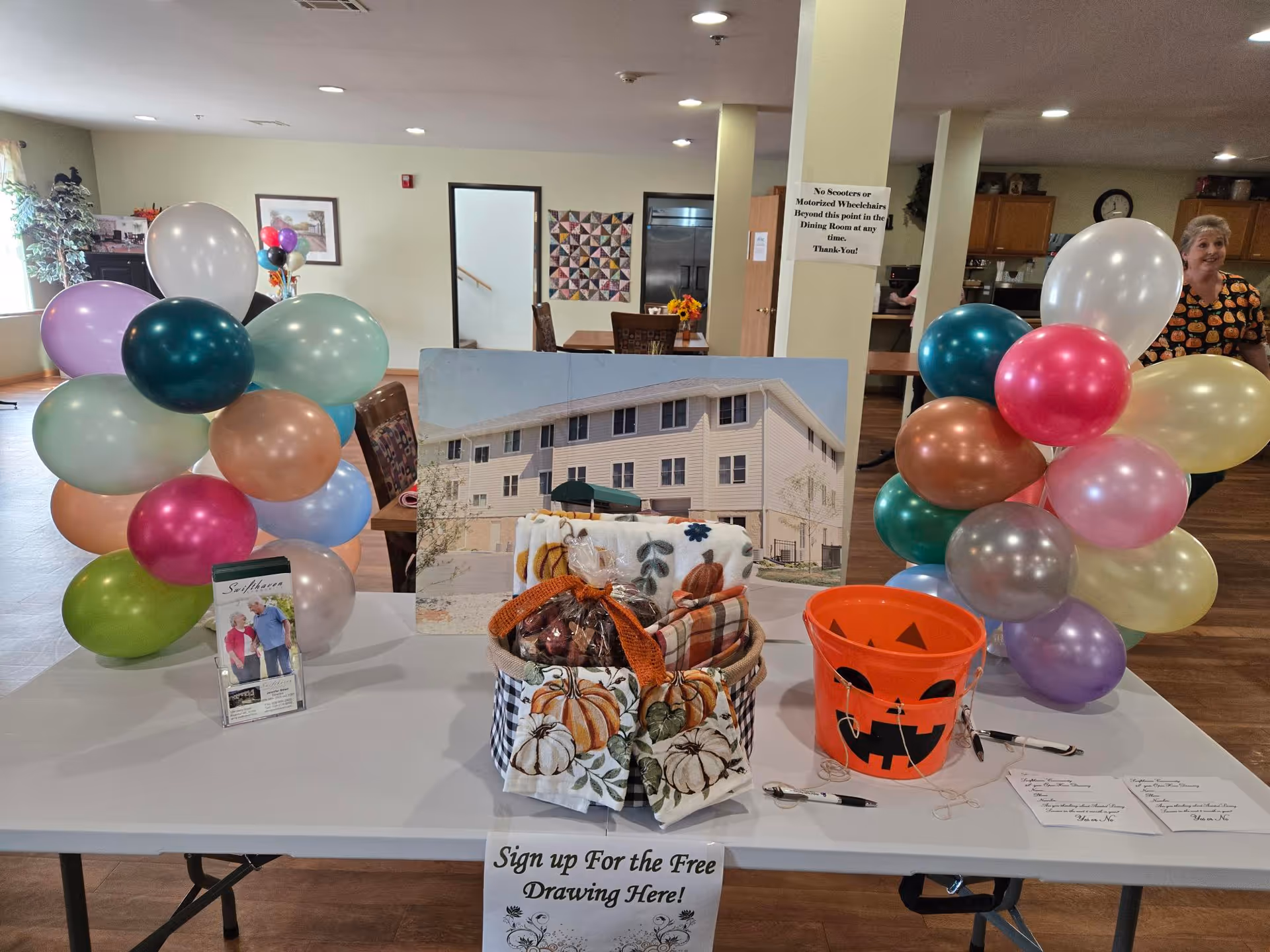 A decorated sign-up table with colorful balloons, a gift basket, and a jack-o'-lantern bucket in a senior living community common room.