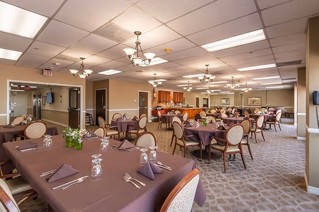 A spacious dining room in a senior living facility with multiple round and rectangular tables covered with brown tablecloths. Each table is set with folded napkins, silverware, and upside-down glasses. The room has beige walls, carpeted floors, and ceiling lights with chandeliers. There is a serving area with wooden cabinets and a countertop in the background.
