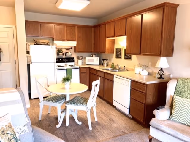 A bright kitchen area with wooden cabinets, white appliances including a refrigerator, stove, microwave, and dishwasher. There is a small round dining table with two chairs in the center, and part of a living room with an armchair and couch is visible on the sides.