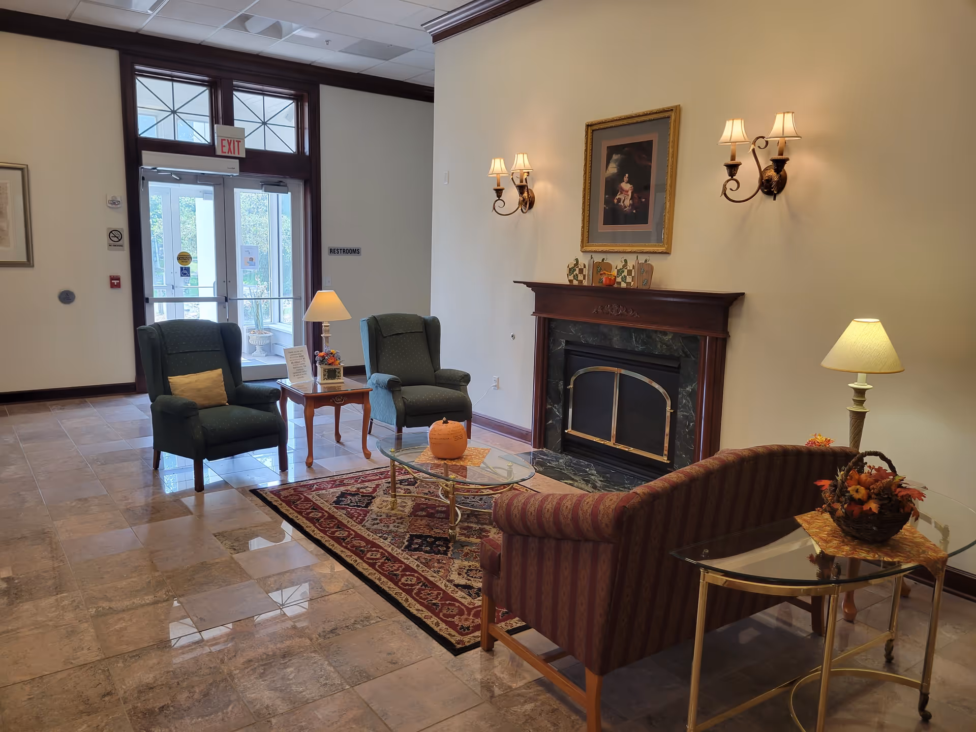 Community lounge with armchairs, a striped sofa, coffee table, fireplace, lamps, and the building entrance.