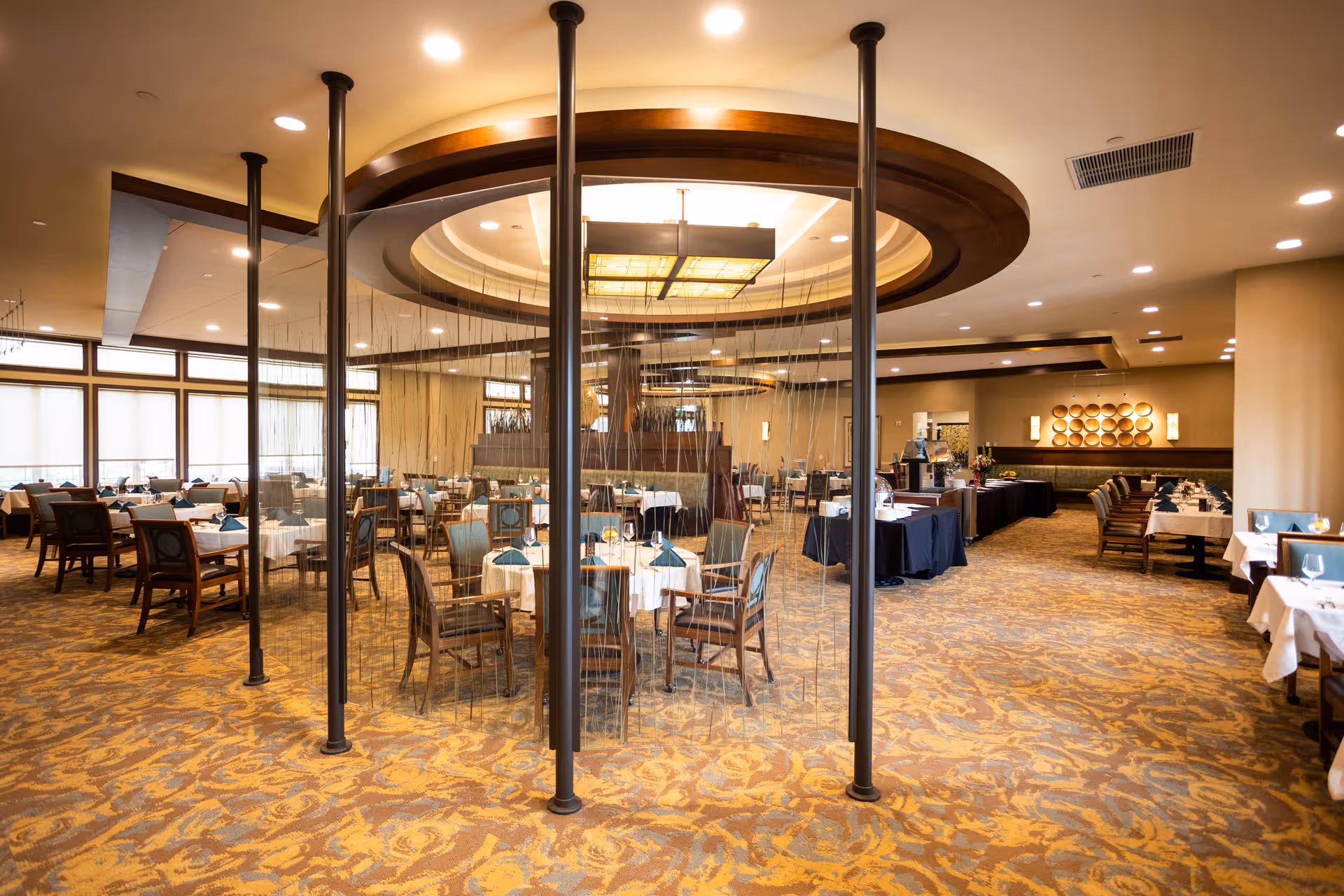 Spacious dining room with multiple tables covered in white tablecloths and set with blue napkins. The room features a patterned carpet, large windows with blinds, and a central decorative circular ceiling fixture with hanging elements. Chairs are arranged neatly around the tables, and buffet tables are set up along the walls.