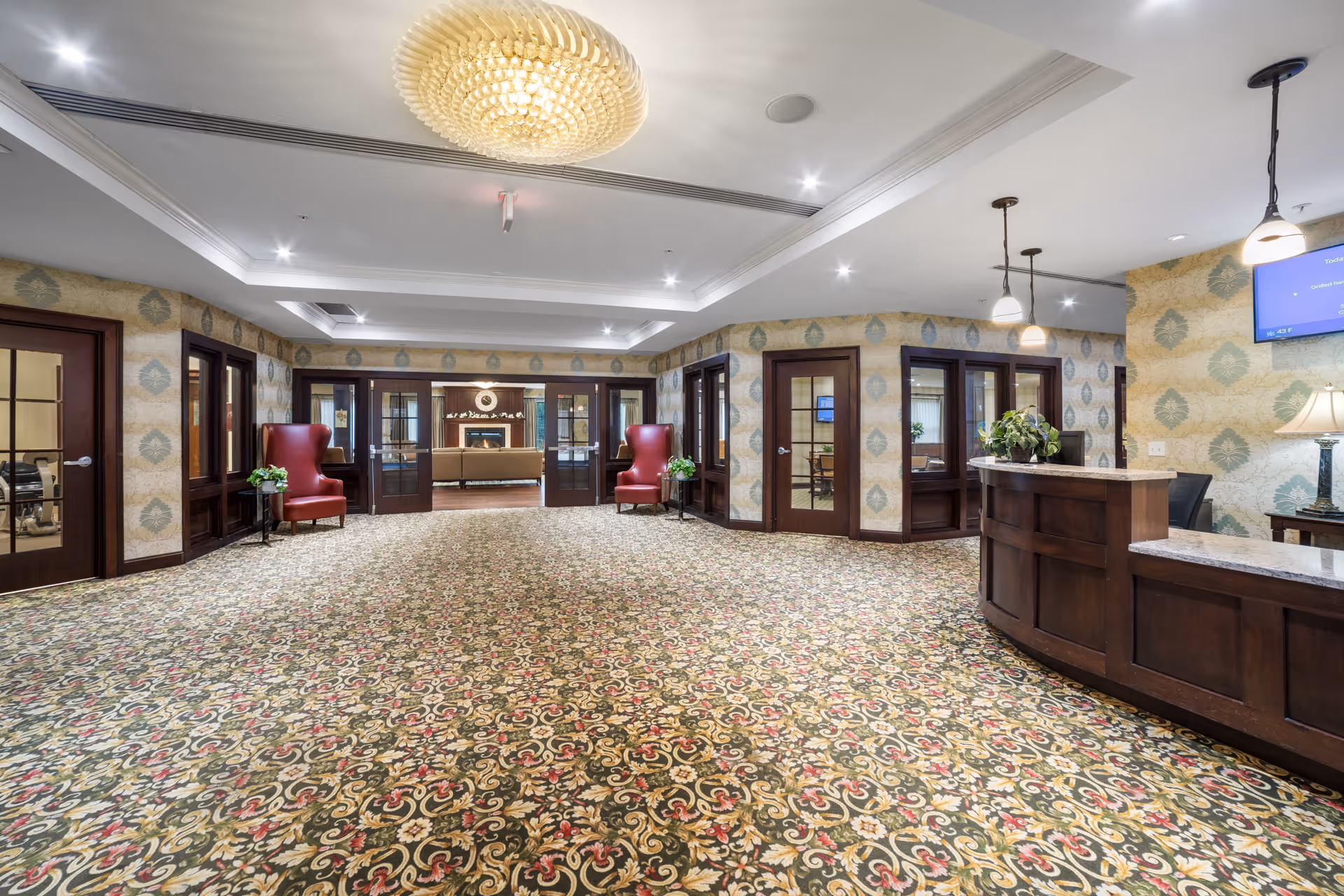 Spacious lobby with patterned carpet, a reception desk on the right, red armchairs, and double doors leading to a seating area.