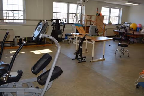 An interior physical therapy/exercise room with stationary exercise equipment, therapy tables, parallel bars and large windows.