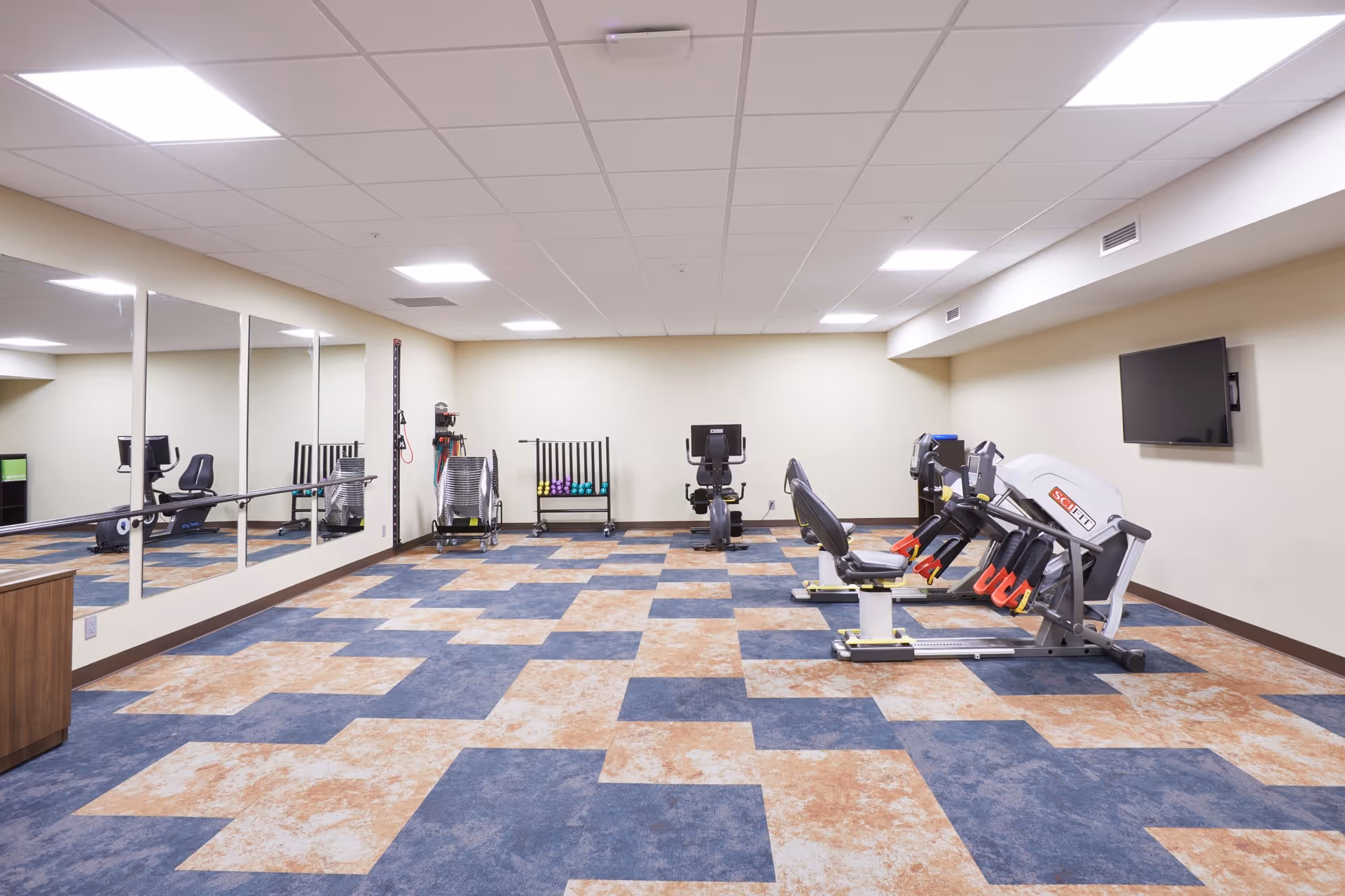 A spacious fitness room with exercise machines, mirrored wall, a TV, and blue-and-tan patterned flooring.
